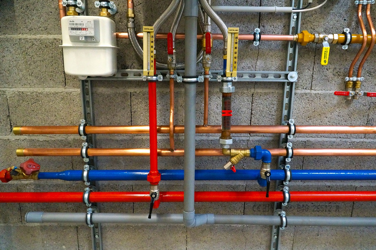 Installation de plomberie et de chauffage sur un mur de parpaings : compteur blanc, tuyaux cuivre, bleus et rouges, vannes et deux thermomètres verticaux.