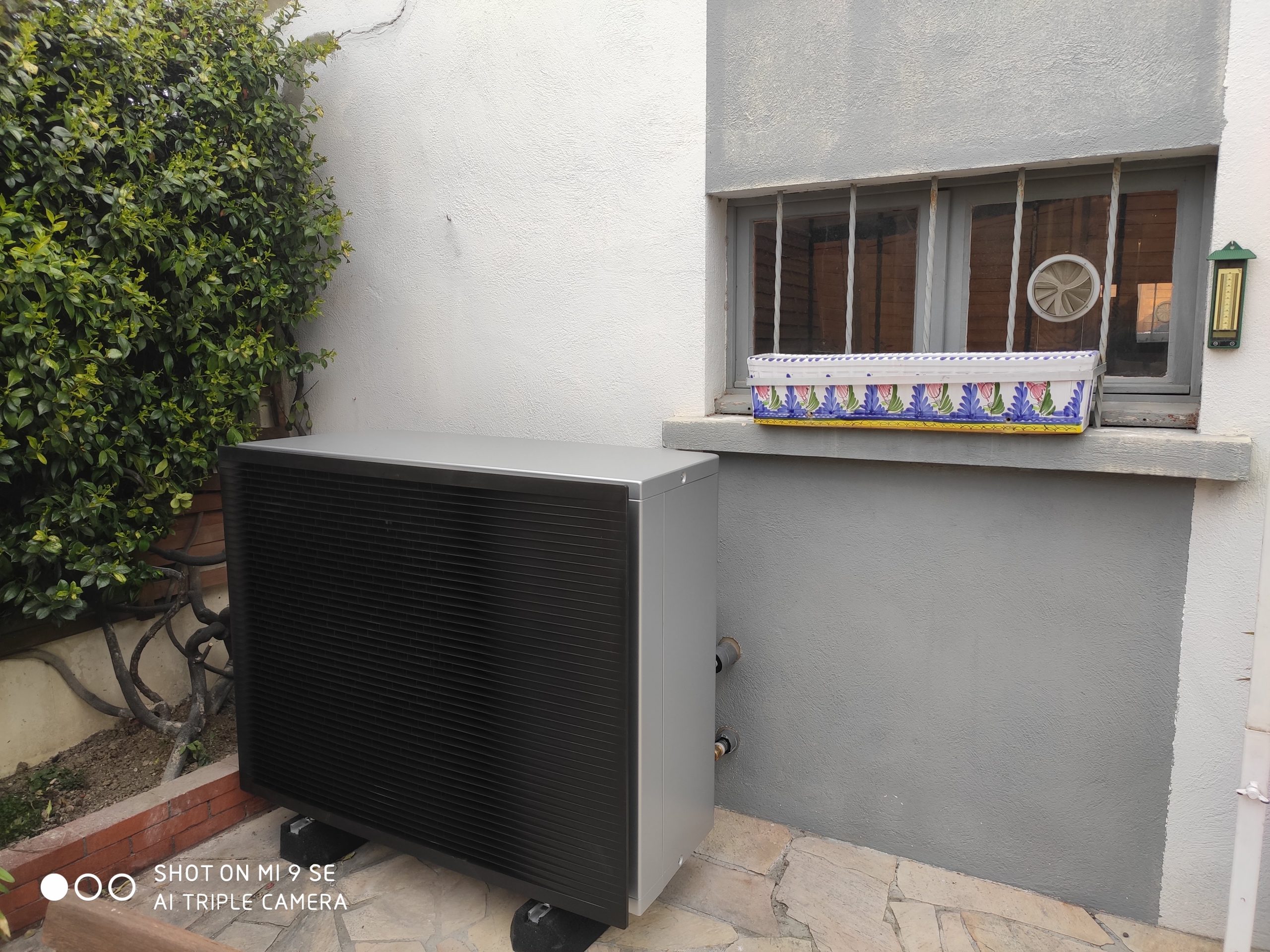 Un appareil gris à grille noire posé contre un mur, sous une fenêtre à barreaux avec un pot de fleurs peint, près d'un buisson.