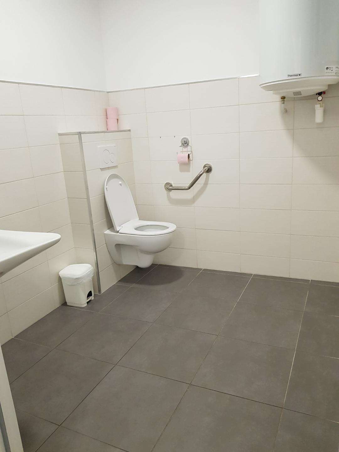Toilettes carrelées de blanc avec WC suspendu, barre d'appui, chauffe-eau et poubelle blanche.